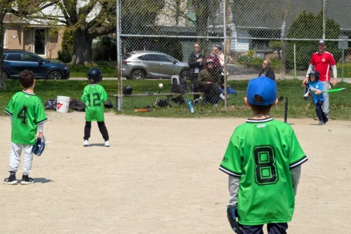 Kids in green jerseys field at a t-ball game as a batter swings.