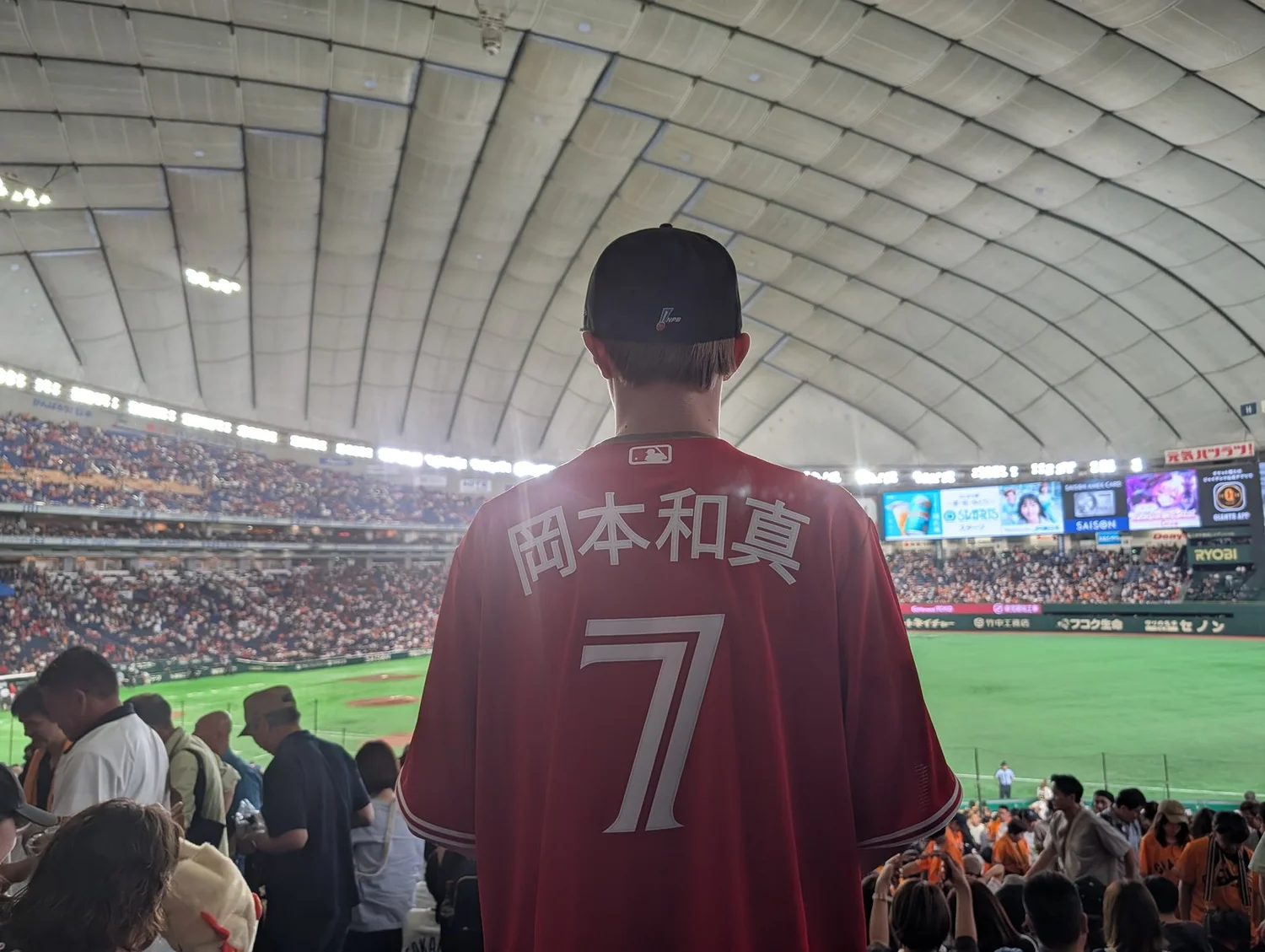A teenager in a red Okamoto 7 jersey seen from behind, looking out over the field at Tokyo Dome