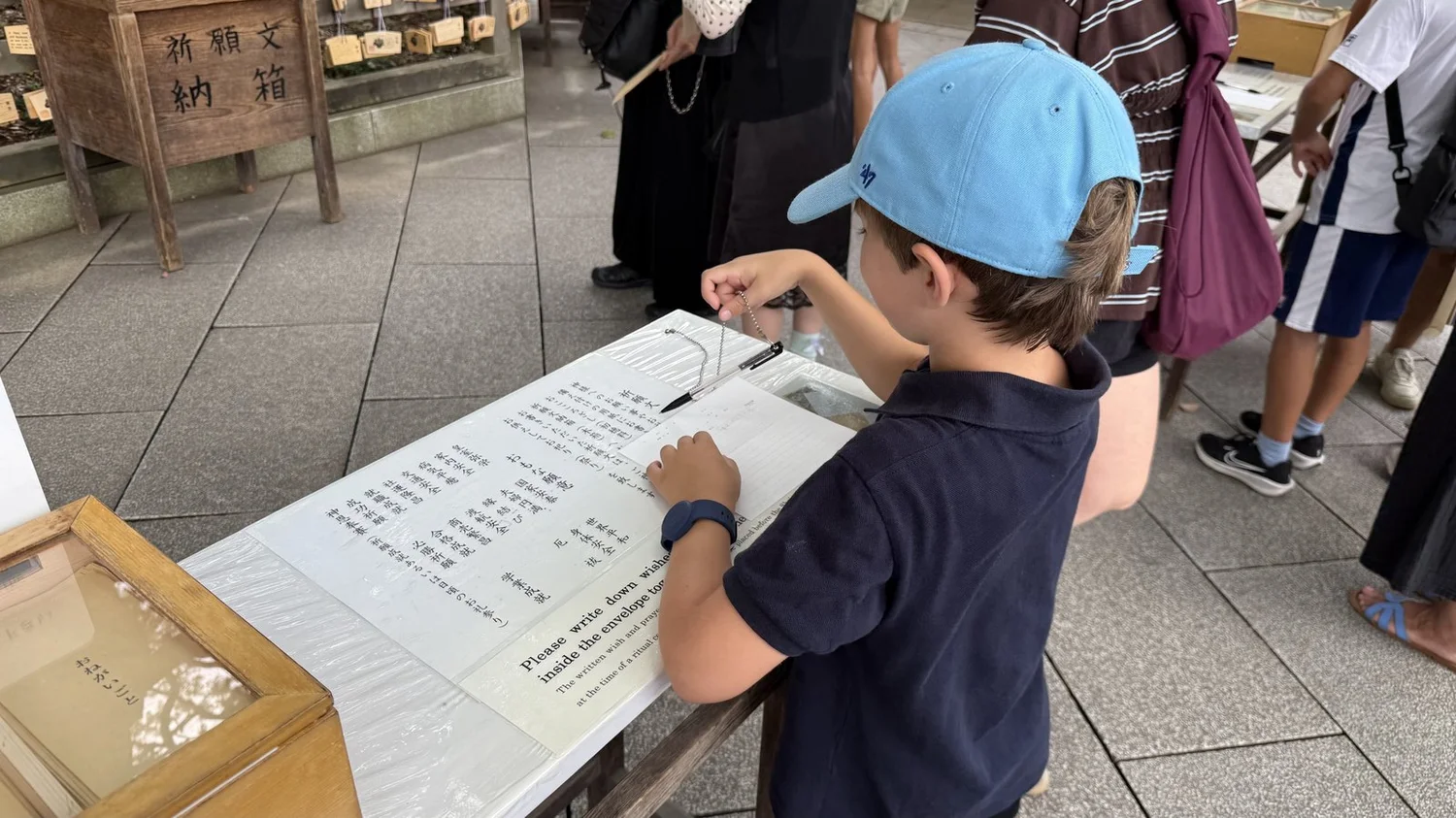 A child in a blue cap, seen from behind, writing at a table at Meiji Jingu beside a sign about sealing wishes in an envelope