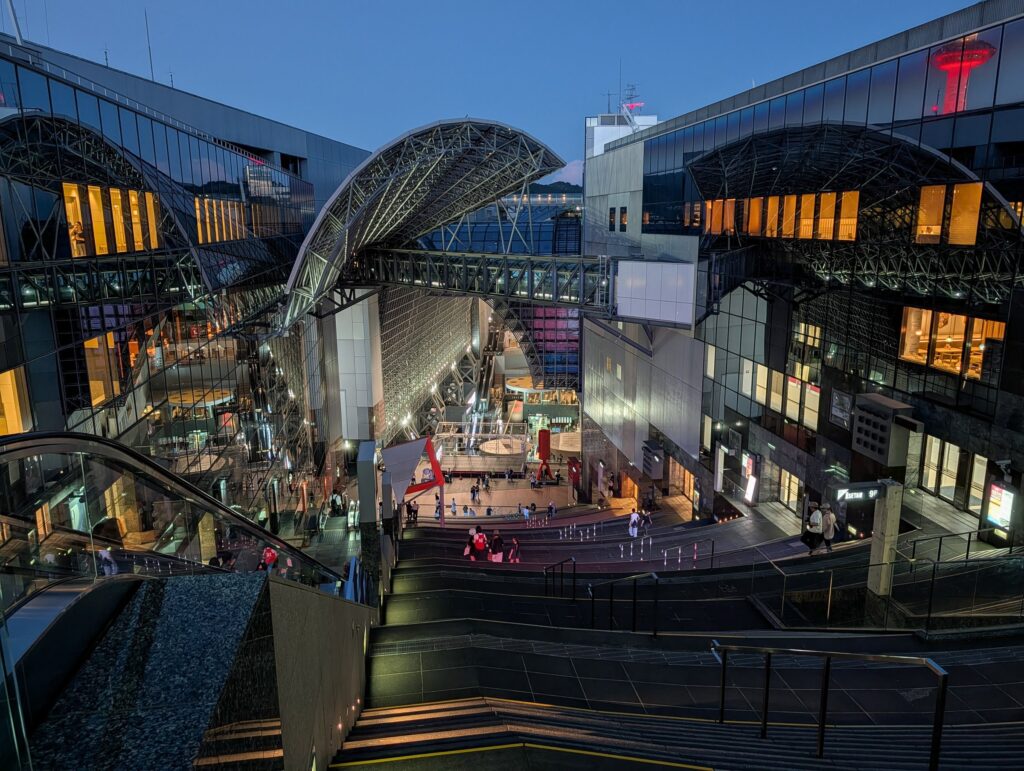 Kyoto Station at dusk, seen from the top of its grand staircase: glass walls and a steel lattice roof, a skyway bridge overhead, people on the steps and plaza below, and Kyoto Tower lit red beyond.