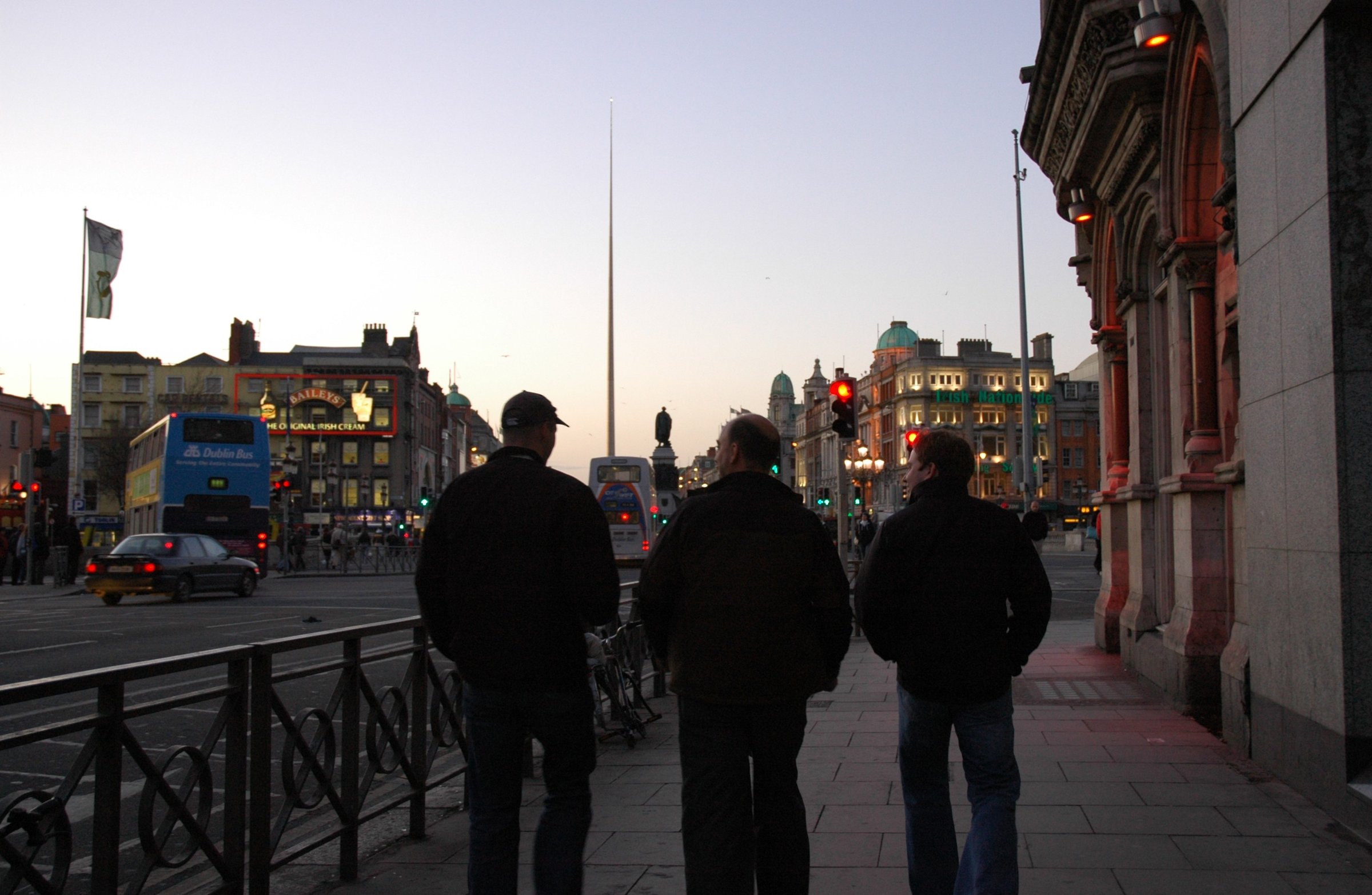 Three men walk along a Dublin street at dusk, seen from behind, with the Spire, a statue and a double-decker bus ahead.