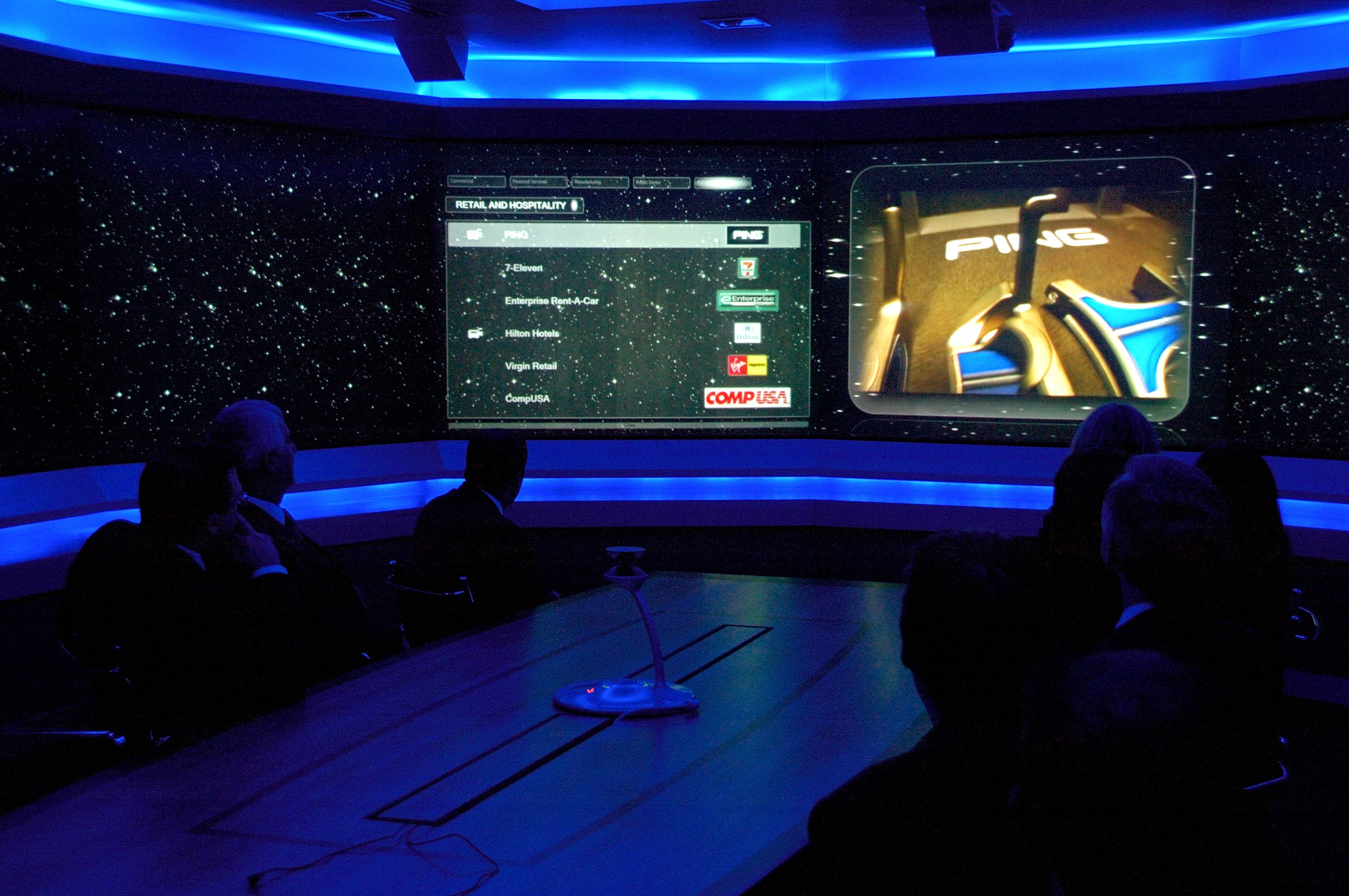 A darkened boardroom with a curved wall of screens showing a starfield, a list of retail companies and a video, while people sit around the table in silhouette.