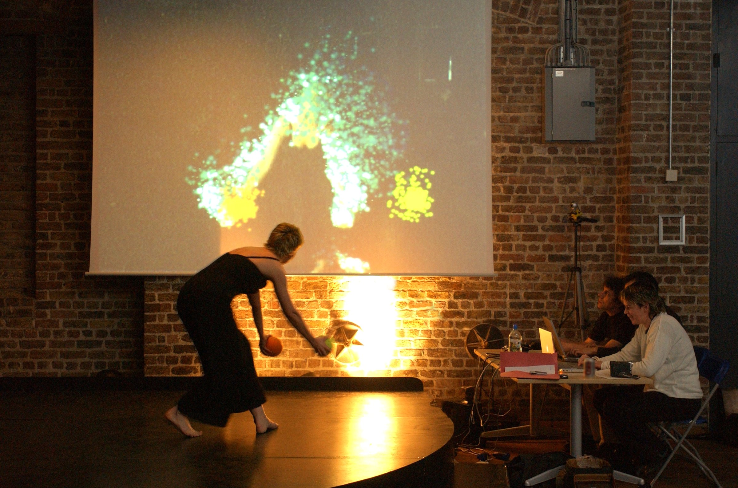 A barefoot dancer in a black dress bends low holding a ball, while trails of coloured light follow her movement on a large screen behind her. Two people watch from a table of laptops at the side.