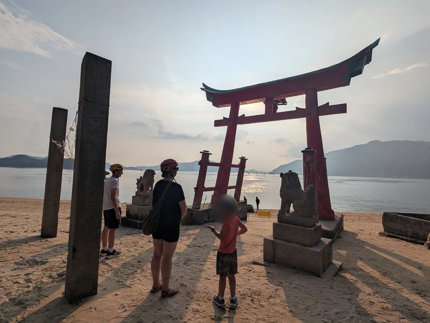 A family at a red torii on a beach on the Shimanami Kaido at golden hour; the youngest's face is blurred