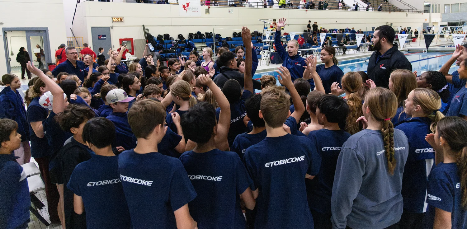 Swimmers in club shirts gathered around a coach with arms raised in a team cheer, seen mostly from behind